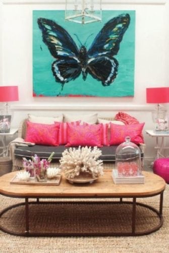 Sala com almofadas rosas no sofá e uma tela turquesa com uma borboleta na parede atrás do sofá.. Contraste .