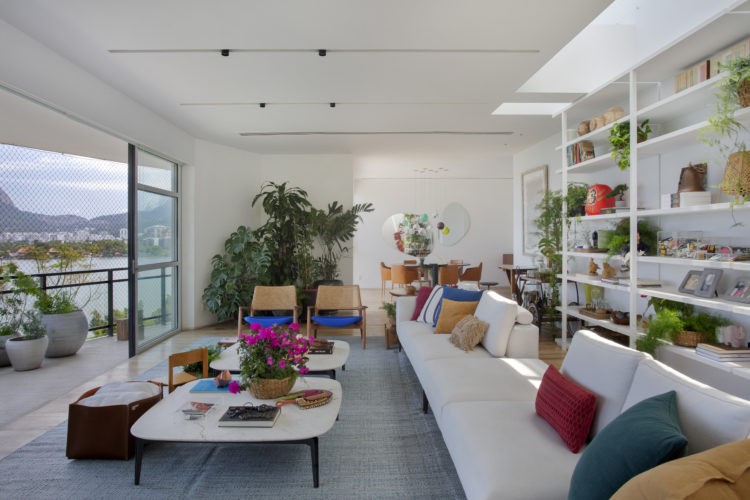 Ampla e clara sala de uma cobertura com varanda. Na sala sofá em L branco e almofadas coloridas