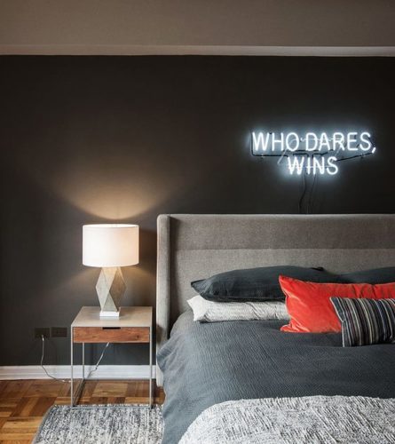 Quarto de casal, com parede pintada na cor cinza escuro e um neon aplicado em cima da cama. No neon a frase em ingles who dares wins