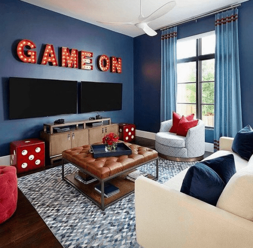 Uma sala decorada com paredes azuis, puff de couro marrom e em cima de duas tvs, letras com micro lâmpadas forma a farse em ingles , game on
