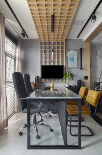 Bancada de escritório, com duas cadeiras pretas e em frente duas cadeiras amarelas. Painel em quadrados de madeira no teto descendo para a parede e uma tv 
