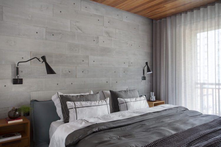 Quarto de casal com parede de fundo da cama revestida com porcelanato imitando cimento, cabeceira e roupa de cama cinza