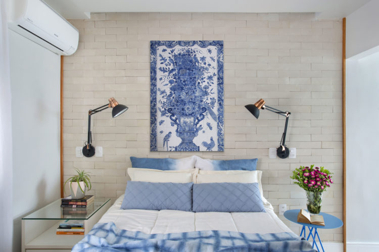 Quarto de casal compacto, parede de tijolinho branca atrás da cama, cama decorada com lençol e almofadas azuis