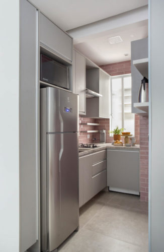 Cozinha pequenas com as paredes revestidas de azulejo retangular na cor rosa, armários na cor cinza e geladeira cromada. 