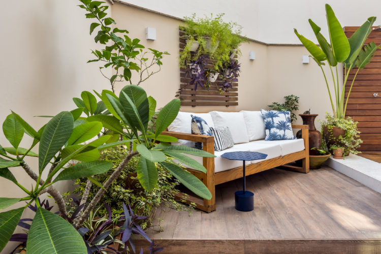 Apartamento térreo com ares de casa. Área externa com sofá em madeira e estofados brancos, almofadas com estampa de coqueiros azuis, e ao lado, vasos de plantas.