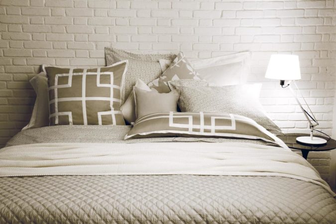 Quarto com parede em tijolinho branco e cama forrada com lençol branco e bege.