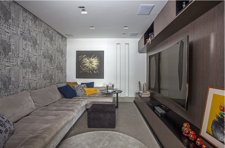 Urbanidade dá o tom para apartamento de família em SP. Sala de tv com sofá cinza a na parede um papel com o sky line da cidade de São Paulo.