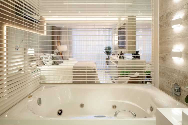 Banheira separada do quarto com vidro e persiana.