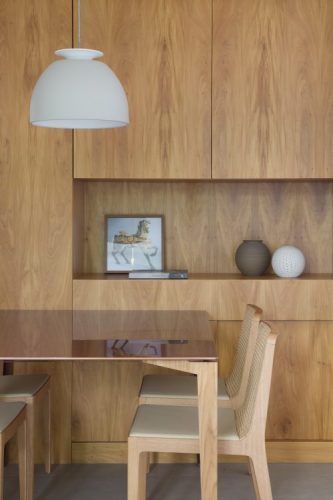 Pequenas interferências para tornar os ambientes mais amplos em um apartamento no Leblon. Parede de fundo da mesa de jantar revestida em madeira, com nicho e portas de armários camufladas