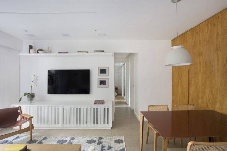 Pequenas interferências para tornar os ambientes mais amplos em um apartamento no Leblon. Parede da Tv branca, rack embaixo com ripas brancas e parede lateral da mesa de jantar revestida em madeira