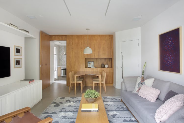 Pequenas interferências para tornar os ambientes mais amplos em um apartamento no Leblon. Sala clara e parede de funda da sala de jantar em madeira, com porta da cozinha camuflada.
