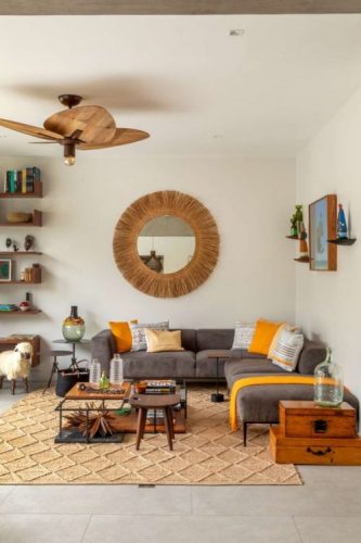 Sala com sofá cinza, almofadas amarelas, ventilador de teto em palha e espelho redondo na parede.