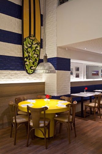 Restaurante com mesa amarela, parede de tijolinho pintado de branco com listras azuis e uma prancha de surf pendurada
