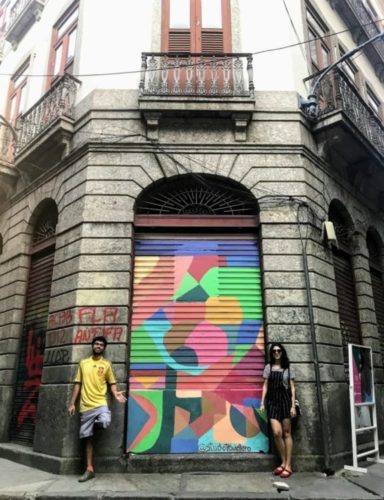 Porta pintada, estilo grafiti,  de um casario antigo no Rio e os artistas ao lado, um homem e uma mulher.