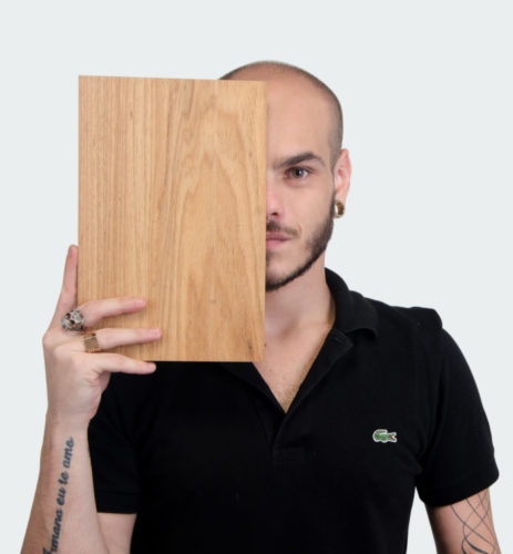 Foto do design Pedro Galaso segurando um pedaço de madeira na frente da metade do rosto.