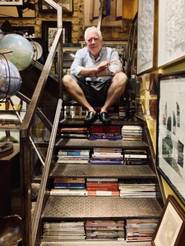 Estante com arte. Foto do arquiteto Chicô Gouvea sentado em uma escada, que entre os degraus estão vários livros
