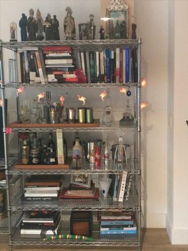 Estante com arte. Foto de uma estante de ferro cromado com livros, garrafas e coleção de santos