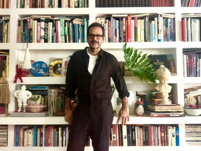 Estantes com arte. Alberto Renault ( com barba e óculos) em frente a uma estante branca, com vários nichos e repleta de livros