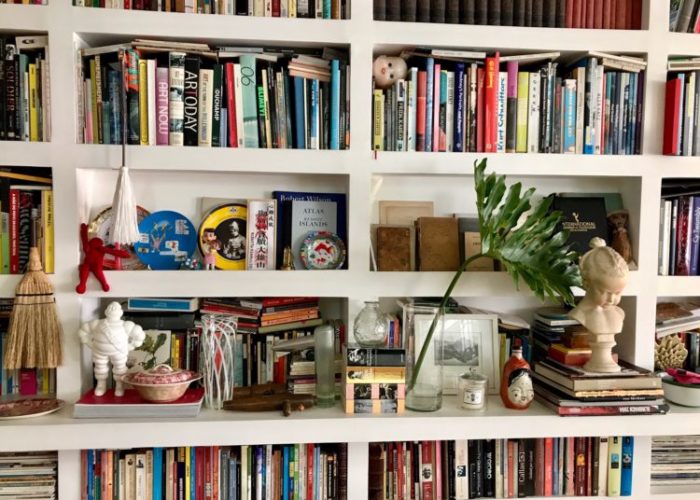 Estantes com arte. Foto de uma estante ocupando uma parede grande. Estante branca, com nichos, repleta de livros e alguns objetos.