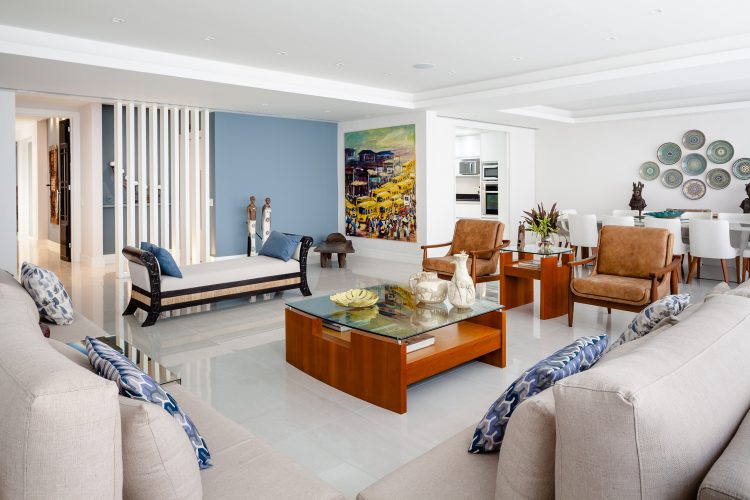 Apartamento da década de 60 passa por modernização. Sala ampla e clara, piso branco, sofás da cor cru, mesa de centro madeira clara e ao fundo uma parede pintada de azul claro