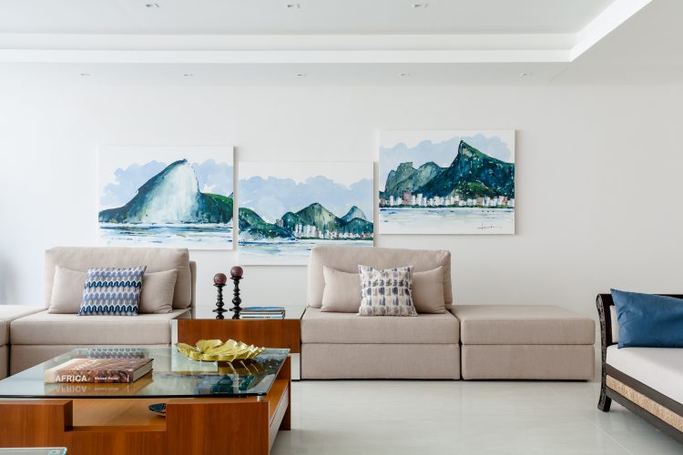 Apartamento da década de 60 passa por modernização. Composição de três quadros em aquarela retratando o rio, pendurados irregularmente na parede branca com um sofá cru em frete.