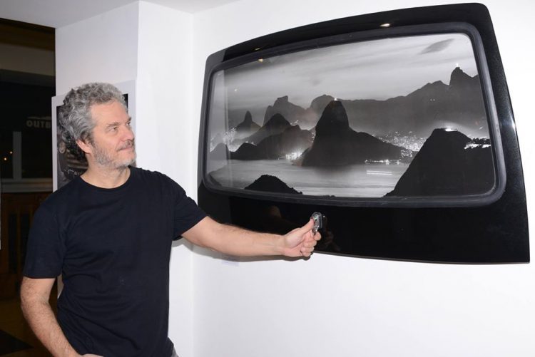 O fotógrafo Joaquim Nabuco, lança a coleção Art Boards, uma série de fotos de paisagens do Rio. Foto do fotografo, blusa preta e barba grisalha segurando uma janela de carro com uma fotografia do Rio impressa