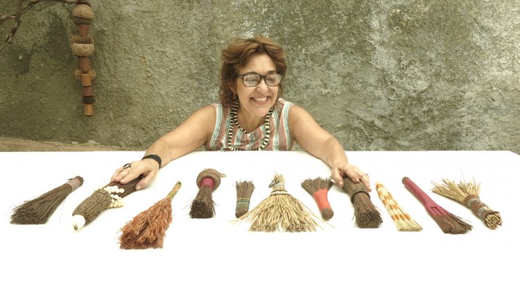 Sementes de sucesso, artista plastica Monica Carvalho que usa sementes no seu trabalho. Foto da artista plástica em frente a uma mesa com varias mini vassourinhas de cipo