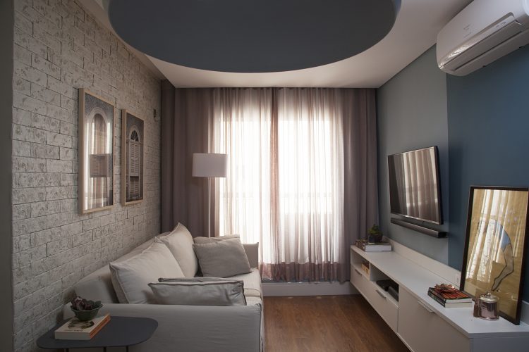 Apartamento de 40 m² com soluções para sua pequena metragem. Sala de estar com parede de tijolinho com sofá branco na frente. Na outra parede, pintada de azul com a tv na parede