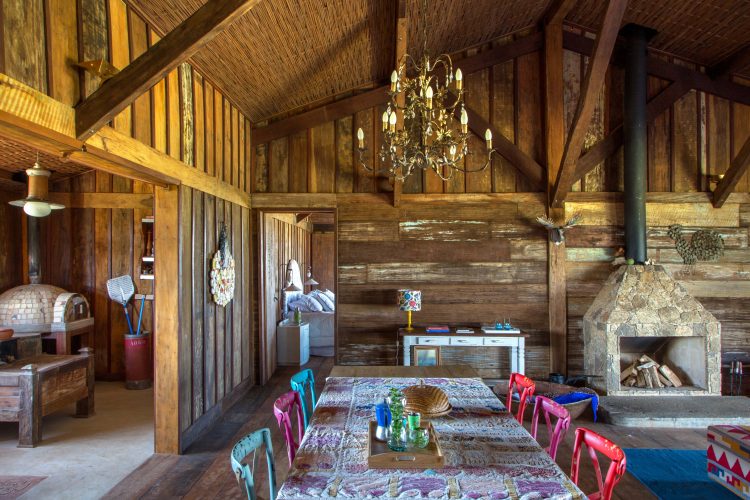 Interior de uma casa no campo. Construção em madeira de demolição, lareira, mesa comprida com cadeiras coloridas