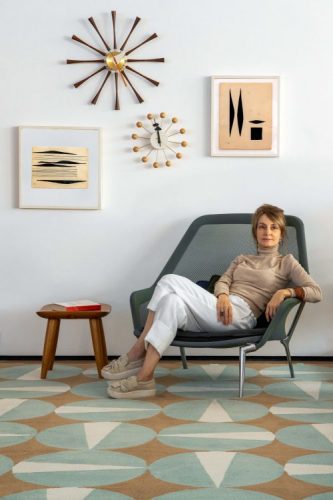 Coleção de tapetes inspirada na obra de Noel Marinho. Mulher sentada em uma cadeira em cima de um tapete com circulos azul claro, peças de design.