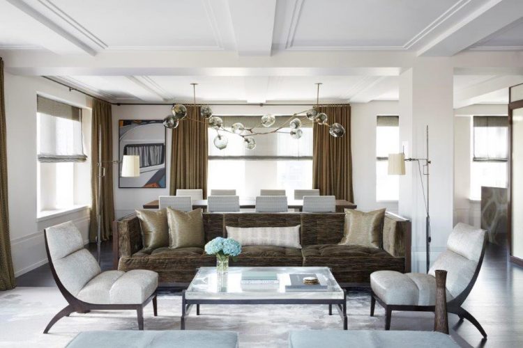 Apartamento com decoração sofisticada em NY. Ampla sala com muitas janelas e sofá marrom com duas poltronas beges