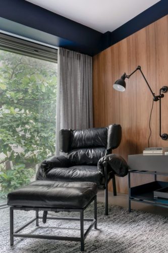 Apartamento no Leblon com décor minimalista e brutalista. Polrona em couro preto ao fundo parede revestida em madeira