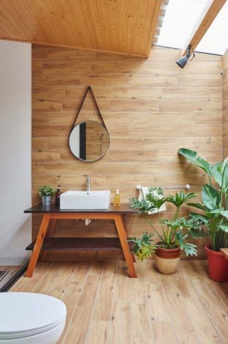 Amplo banheiro revestido com porcelanato imitando ripas de madeira no piso e subindo pelas paredes. Bancada com movel e espelho adnet redondo na parede, plantas ao lado
