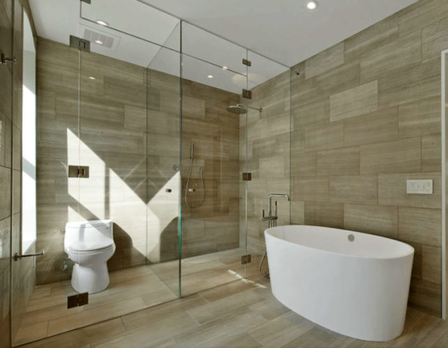 Banheiro grande e claro. Com piso e parede com ao mesmo revestimento imitando madeira, banheira branca e vidro separando a area do box e vaso sanitario