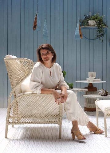  Varanda DonaFlor com projeto da arquiteta Juliana Pippi/ Mulher vestida de roupa clara sentada em um poltrona de fibra clara, parede azul ripada de madeira