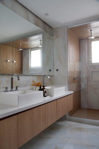 Banheiro com piso em porcelanto imitando pedra ônix, bancada branca e armaruos em madeira