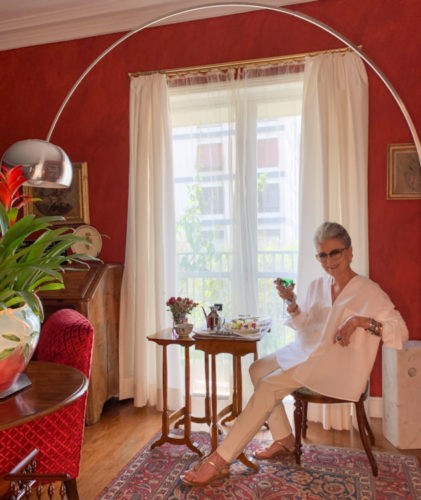 Westwing convida Costanza Pascolato para assinar curadoria. Costanza vestida de branco tomado chá na sua sala com paredes vermelhas