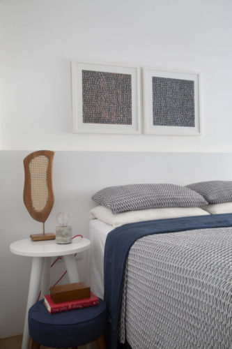 Quarto com parede branca ao fundo da cama, painel como cabeceira , na cor bem clarinha e uma mesa redonda como cabeceira