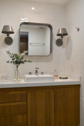 Banheiro com aramrios em madeira, bancada braca e duas arandelas em metal ao lado do espelho