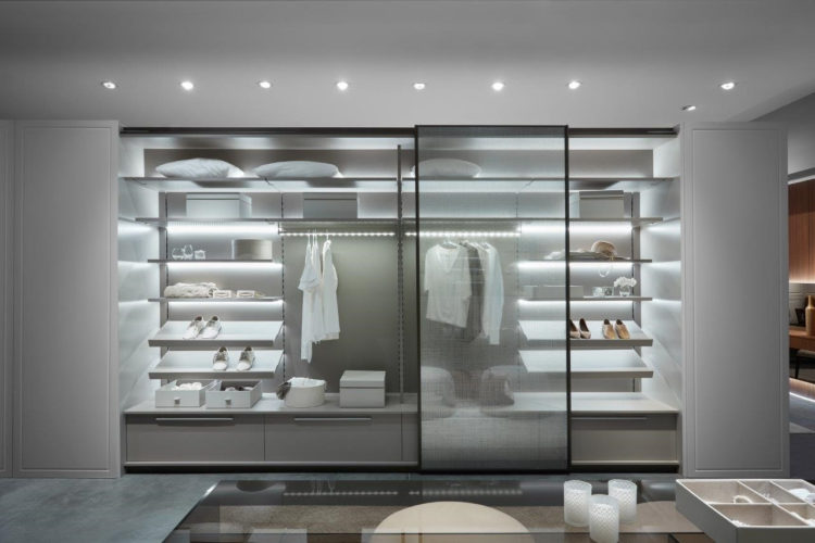 Armario em um closet com portas transparentes em vidro, prateleras brancas iluminadas