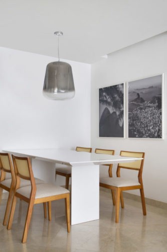 Cantinho da sala de jantar, com mesa branca, cadeiras em madeira , luminaria pendente cromoda e duas fotografias em preto e branco na parede.
