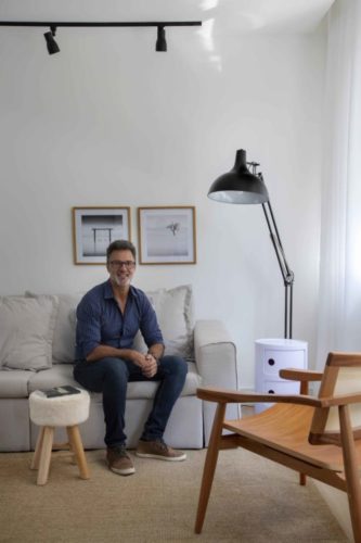 O arquiteto Nilton Montarroyos, sentado no sofá bege e ao lado uma luminaria de pé preta