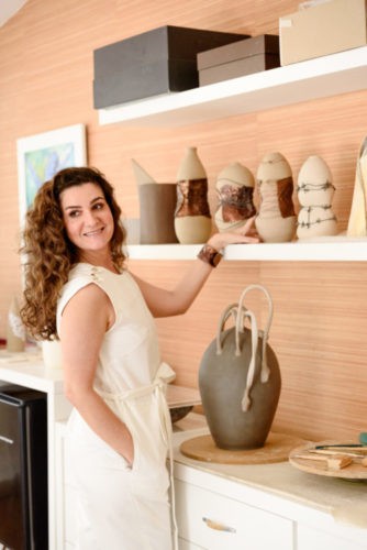 Foto da artista plástica Patricia Guereiro, vestida com um vestido bege e colocando a mão nas suas cerâmicas