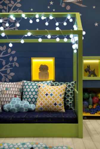 Quarto de criança bem colorido, cama baixa na cor verde em forma de casinha e fio de luzes em volta. Parede de fundo azil e nichos verdes iluminados
