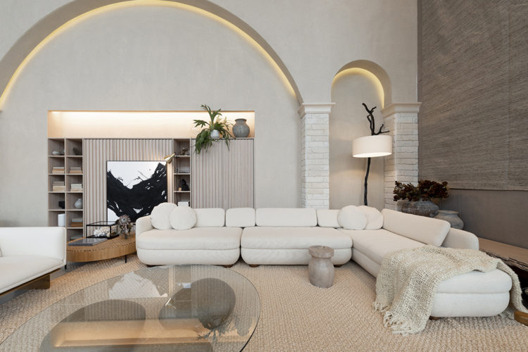 Ambientes abordam o novo morar no Janelas CASACOR /SC. Sala ampla e clara, comum grande sofá em L branco, e atrás arcos na parede 