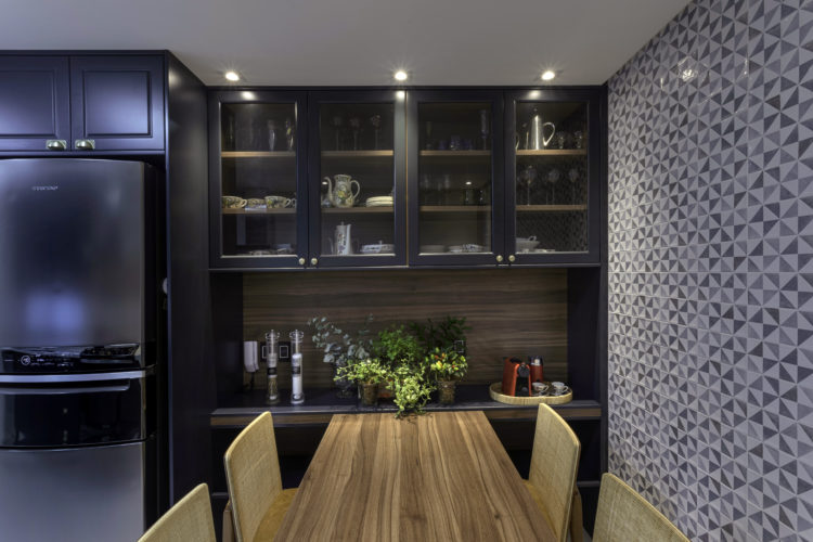 Cozinha decorada, parede revestida com azulejos decorados, armario em azul escuro com portas de vidro, embaixo uma bancada que sai uma mesa em madeira.