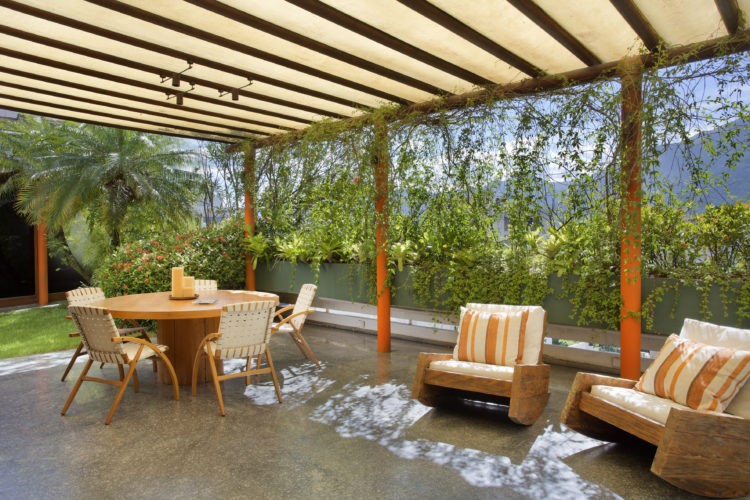 Terraço com pergolado em madeira, piso em granito, caeiras de design e uma mesa redonda. Ao fundo floreiras 