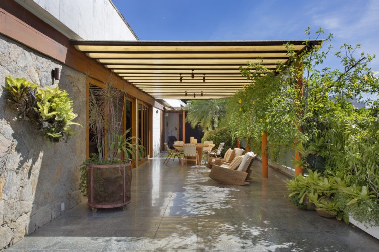 Terraço em uma cobertura linear com cara de jardim de casa. Parte coberta com pergolado em madeira, piso em granito e floreiras em frente a porta da sala