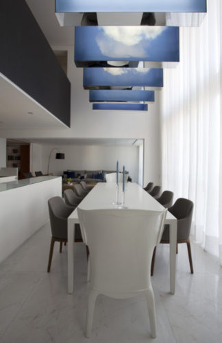 Espaço para a mesa de jantar com pé direito alto, mesa branca e em cima a obra de arte de Eduardo Coimbra, painies iliminados com fot de nuvem