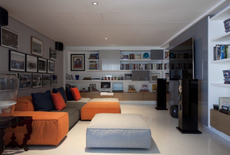 Home tv com sofá em modulos coloridos nas cores cinza, laranja e branco. Quadros de fotos na parede atras e em frente uma tv grande na parede